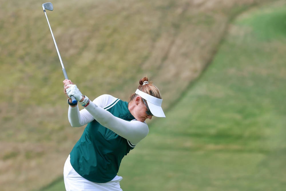 Golfer in green top, white visor and white sleeves mid-swing with an iron on a grassy course