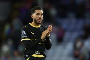 Footballer in black and gold Manchester City kit with Premier League badge applauding fans at a night match