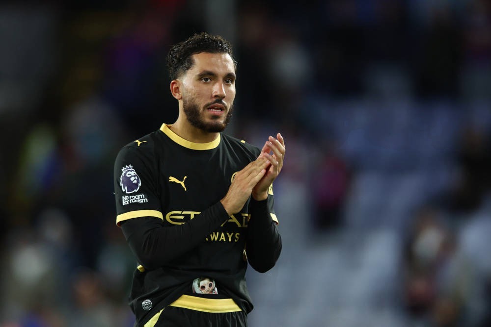 Footballer in black and gold Manchester City kit with Premier League badge applauding fans at a night match