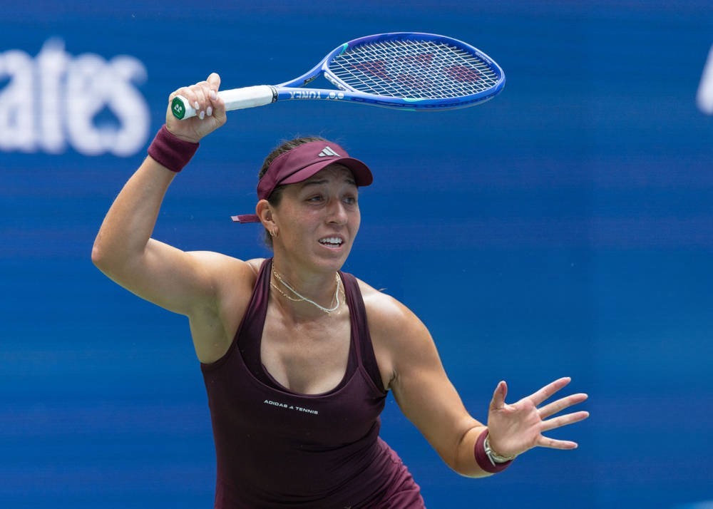 Female tennis player in maroon visor and sleeveless top, swinging a blue Yonex racquet on a blue court at the US Open