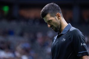 Novak Djokovic in a dark polo with a grimacing expression, looking down during a match at a blurred indoor tennis arena.