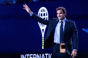 A man in a dark suit and blue shirt waves on stage in front of a dark blue backdrop with a tennis racket logo, at an event with "INTERNATIO" text visible.