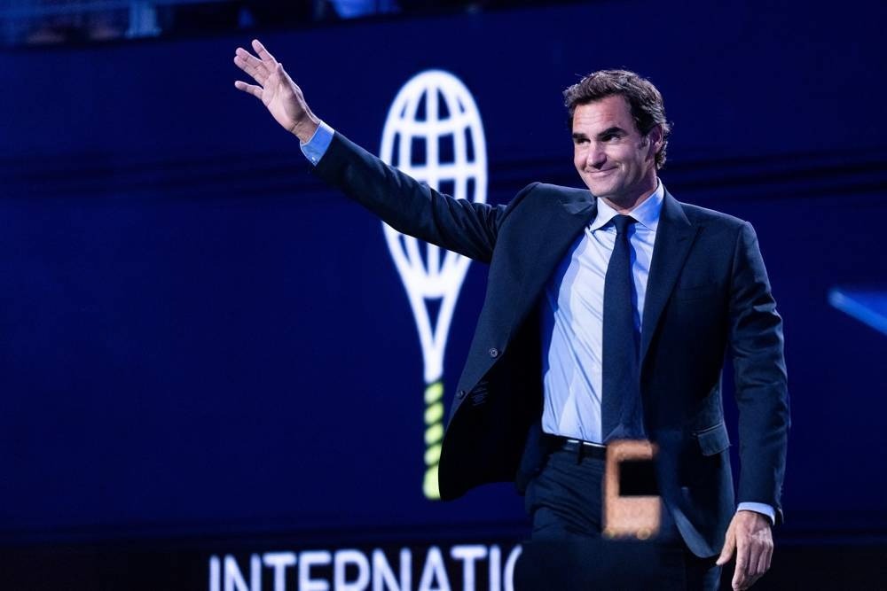 A man in a dark suit and blue shirt waves on stage in front of a dark blue backdrop with a tennis racket logo, at an event with "INTERNATIO" text visible.