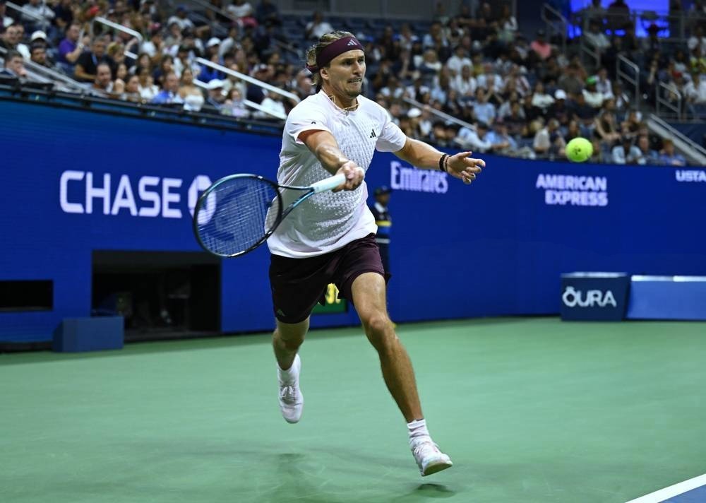 Tennis player in white Adidas shirt and maroon shorts reaching for a forehand on the court at the US Open, with Chase and Emirates signage behind him.