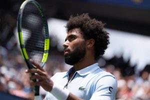 A tennis player in a light blue Lacoste polo and white wristband holding a green-and-yellow racket at a US Open match, crowd blurred behind him.