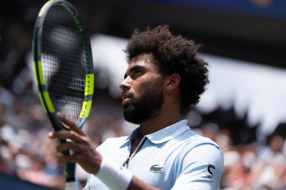 A tennis player in a light blue Lacoste polo and white wristband holding a green-and-yellow racket at a US Open match, crowd blurred behind him.