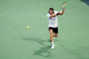 Tennis player in white shirt and black shorts, Wilson racket, hitting a forehand on a green court at the US Open.
