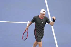 A tennis player in a black shirt and white cap pumps his fist on a blue court, holding a red racquet, at the US Open.