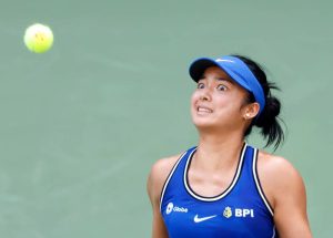 Tennis player in blue Nike visor and BPI/Globe top, wide-eyed, tracking a yellow ball in the air on a green court.