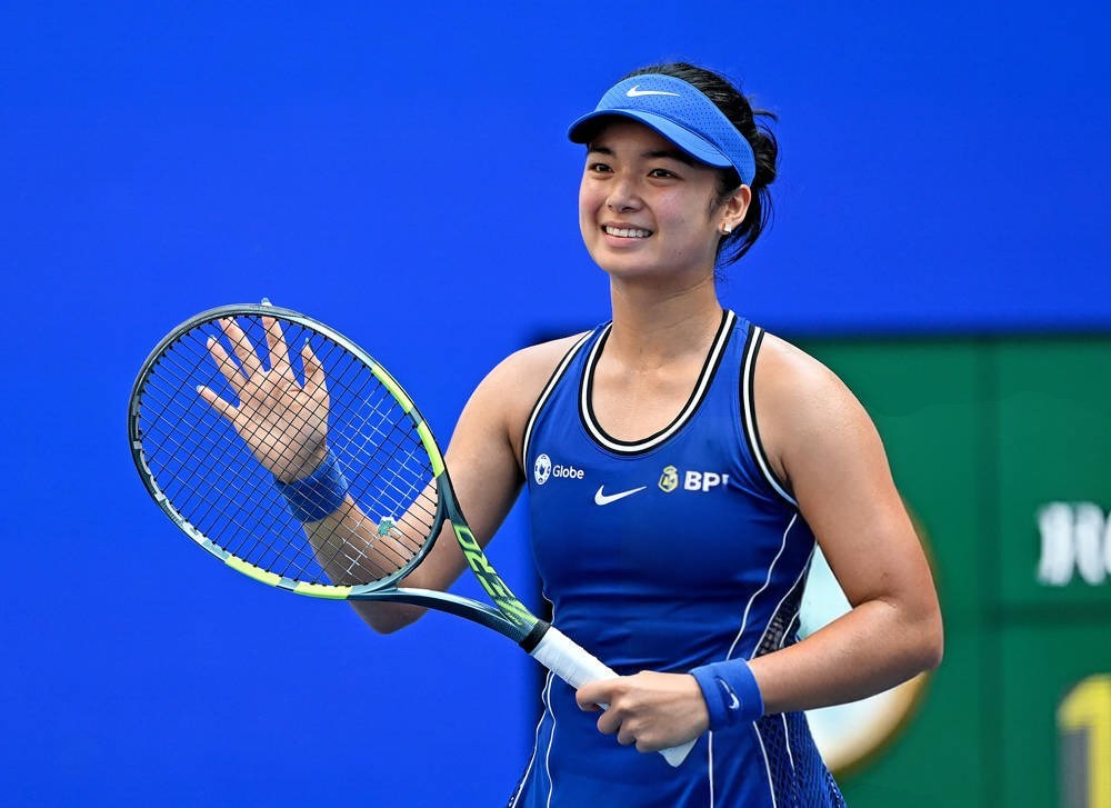 Tennis player in blue Nike visor and sleeveless top, with BPI and Globe logos, smiling and waving her racket on a blue court.