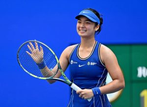 Tennis player in blue Nike visor and sleeveless top, with BPI and Globe logos, smiling and waving her racket on a blue court.