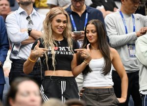 Two women smiling in a stadium crowd, one in a "I ♥ MY BOYFRIEND" crop top, both pointing at their rings; a lanyard-wearing crowd behind them.