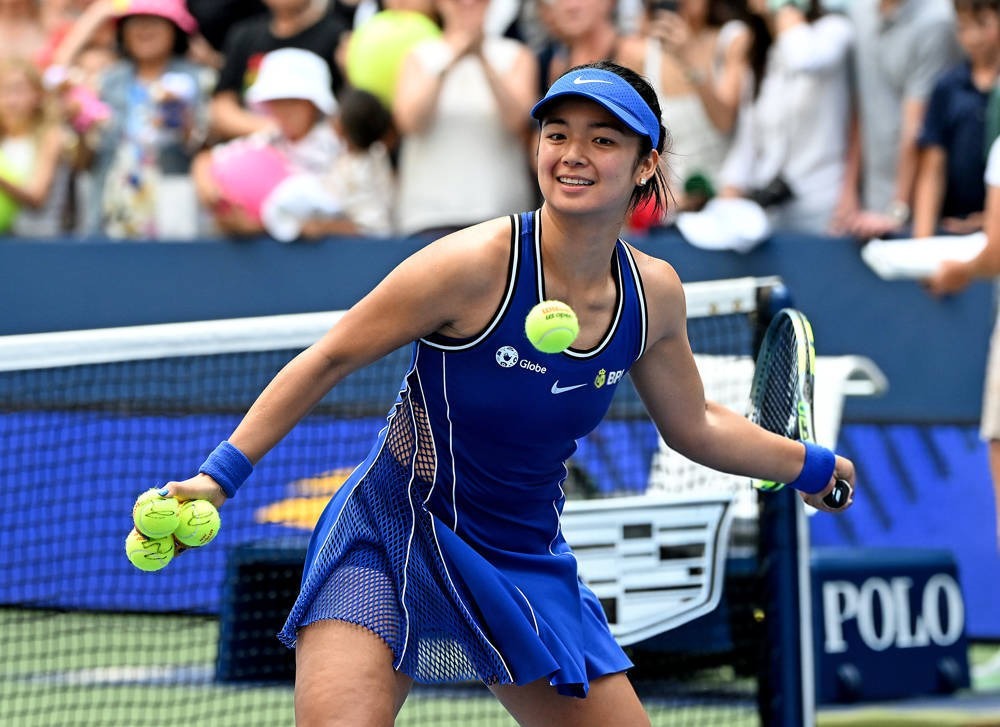 Tennis player in a blue Nike dress and visor, smiling and holding tennis balls on a court, with a crowd behind the net at a match.