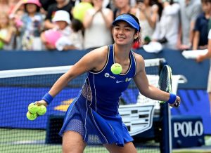 Tennis player in a blue Nike dress and visor, smiling and holding tennis balls on a court, with a crowd behind the net at a match.
