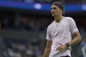 Tennis player in a pink Adidas shirt and purple headband, with a gold chain and dark watch, looking on during a US Open match.