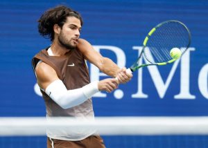 Tennis player in a brown Nike sleeveless top and white arm sleeve hits a backhand on court at the US Open, blue backdrop behind him.