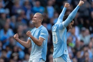 Blond footballer in light blue Manchester City kit celebrating with clenched fists in a packed stadium, teammate with arms raised behind him.