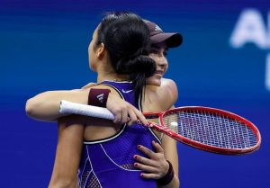 Two tennis players in a tight embrace on court, one in a purple sleeveless top holding a red racket, the other in a maroon Adidas cap.