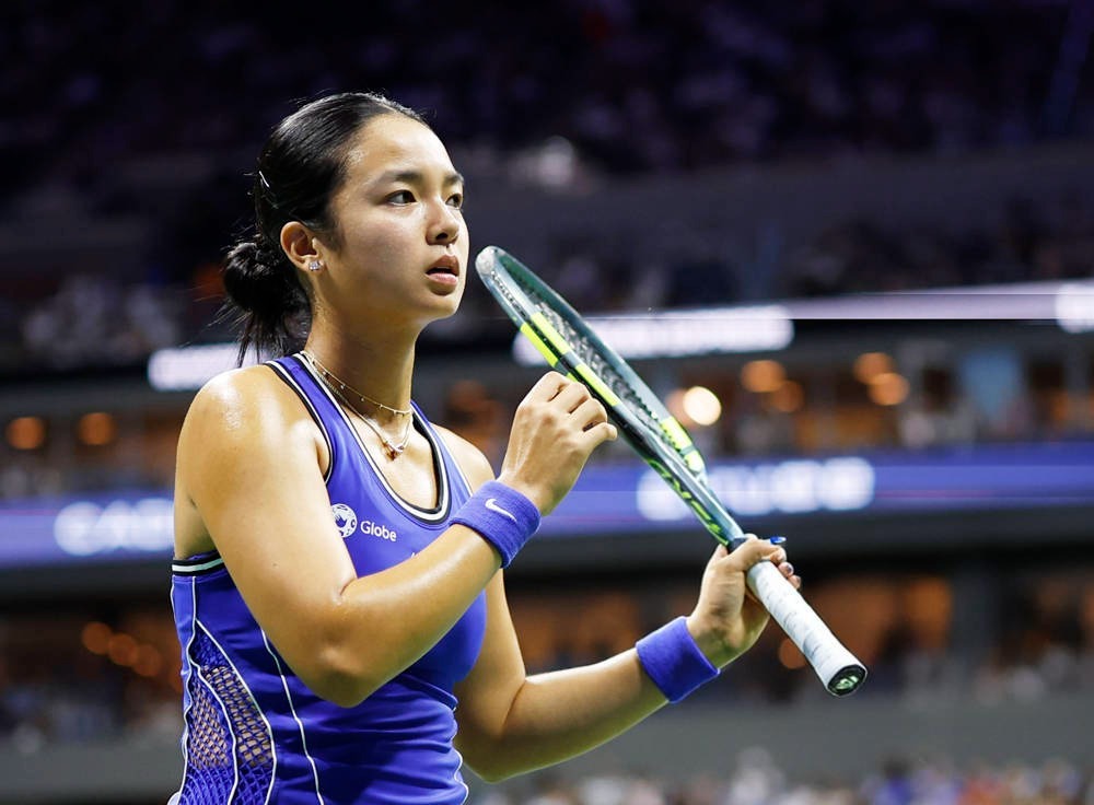Alex Eala in a blue sleeveless tennis top holding her racket on court at a night match, with stadium lights behind her