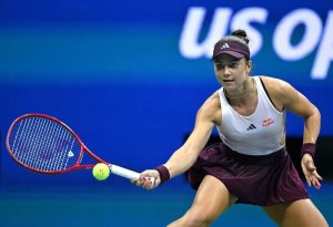 Tennis player in a maroon visor, skirt and white Adidas top with Red Bull logo, reaching for a low forehand at the US Open, with the "us open" sign behind her.