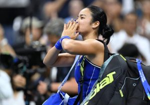 Tennis player in a blue outfit and blue wristbands blows a kiss toward the crowd, a tennis bag with Babolat logo on her back, cameras behind her