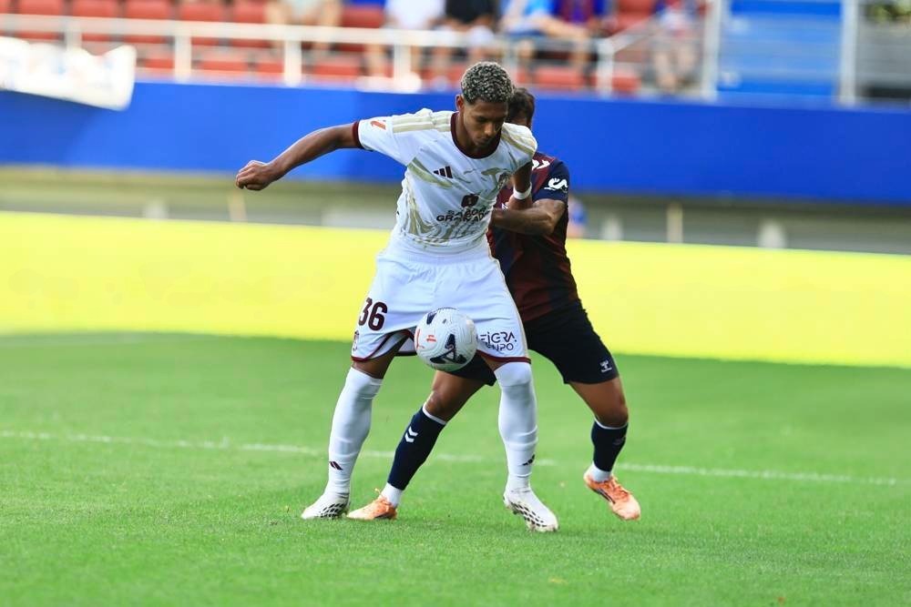 Footballer in white kit with number 36 battling for the ball against a dark red and navy opponent on a green pitch