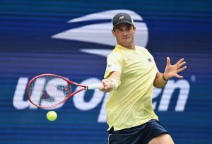 Tommy Paul hits a forehand in a yellow shirt and navy shorts at the US Open, with the tournament logo behind him.