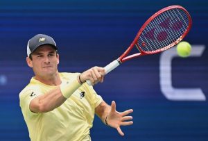 A tennis player in a yellow New Balance shirt and cap hits a forehand with a red Yonex racquet, ball in the air at a hard court event.