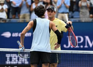 Two tennis players shaking hands over the net at the US Open, one in a light blue mesh tank top and one in a yellow shirt and cap holding a red racket.