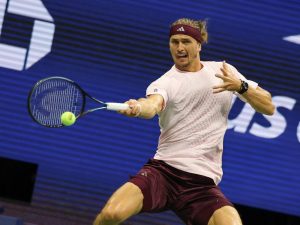 Tennis player in maroon headband and shorts hitting a forehand at the US Open, with a blue backdrop behind him