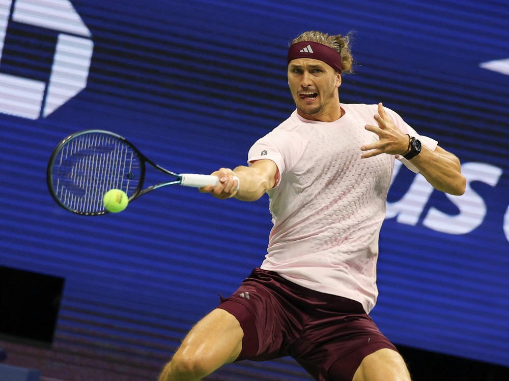 Tennis player in maroon headband and shorts hitting a forehand at the US Open, with a blue backdrop behind him