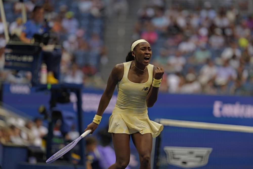 Tennis player in a pale yellow New Balance dress shouting and clenching her fist mid-match at the US Open, with a racket in hand