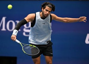 Tennis player in a light blue Nike mesh tank and black shorts, swinging a racket to hit a yellow ball on a blue court