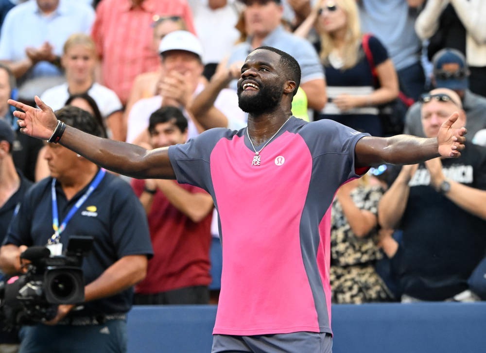 Tennis player in a pink and grey shirt with arms outstretched and a big smile, celebrating in a stadium crowd at the US Open.