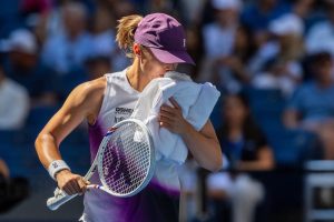 Tennis player in purple cap and white tank top with a racket, wiping her face with a white towel at a US Open match