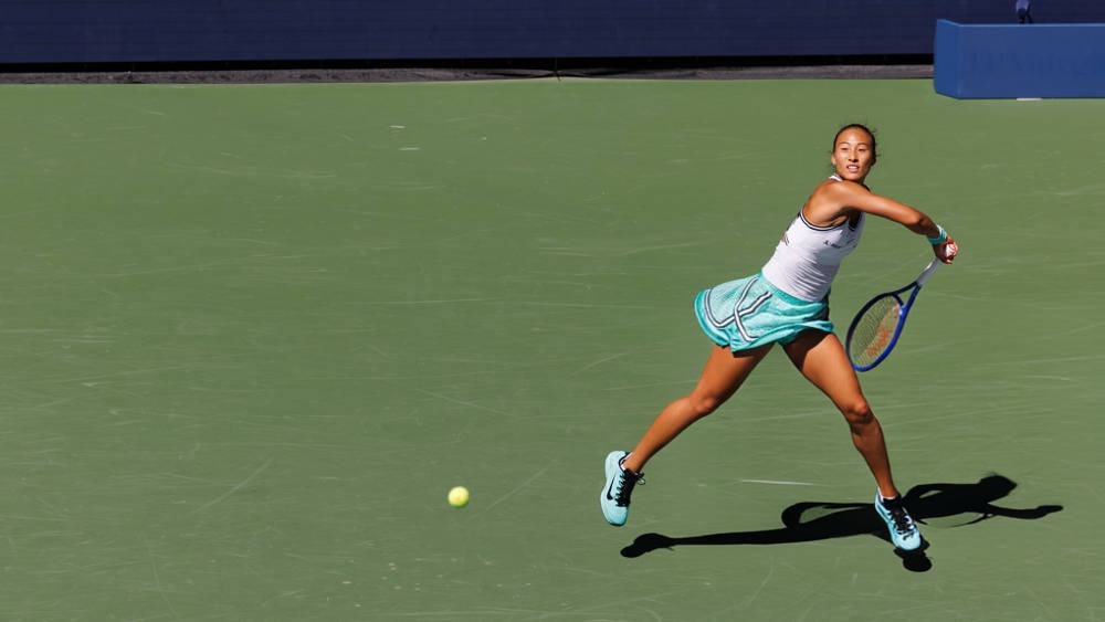 A tennis player in a white top and teal skirt swings a forehand on a green hard court at the US Open.