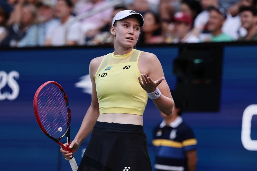 Tennis player in a yellow Yonex top and black skirt, holding a red racket and gesturing with an open hand during a US Open match.