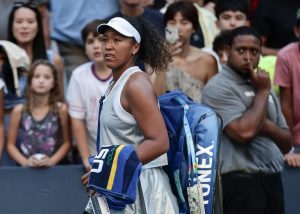 Tennis player in white cap and grey tank top carrying a blue Head/Wilson-style racket bag and towel, walking off court at a US Open match.