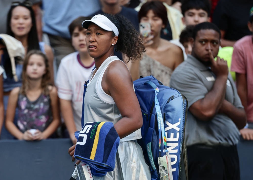 Tennis player in white cap and grey tank top carrying a blue Head/Wilson-style racket bag and towel, walking off court at a US Open match.