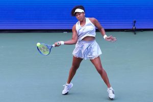 Tennis player in white Nike visor, top and pleated skirt, swinging a blue racquet to return a ball on a green court at the US Open.
