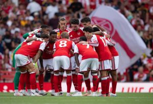 Arsenal players in red and white kits form a huddle on the pitch before a match, with fans and a club flag behind them