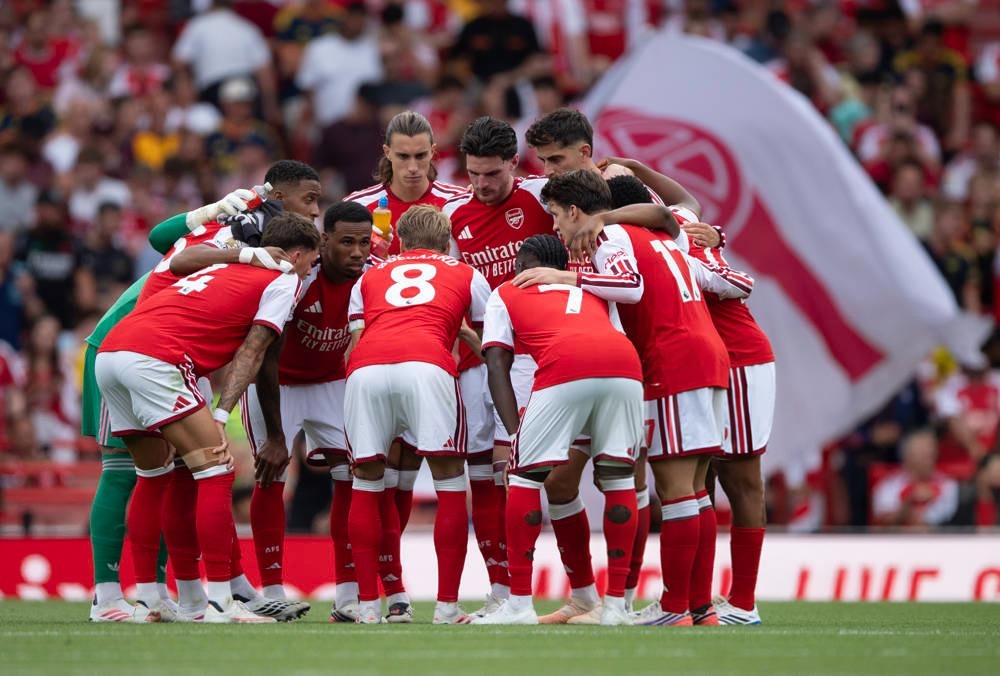 Arsenal players in red and white kits form a huddle on the pitch before a match, with fans and a club flag behind them