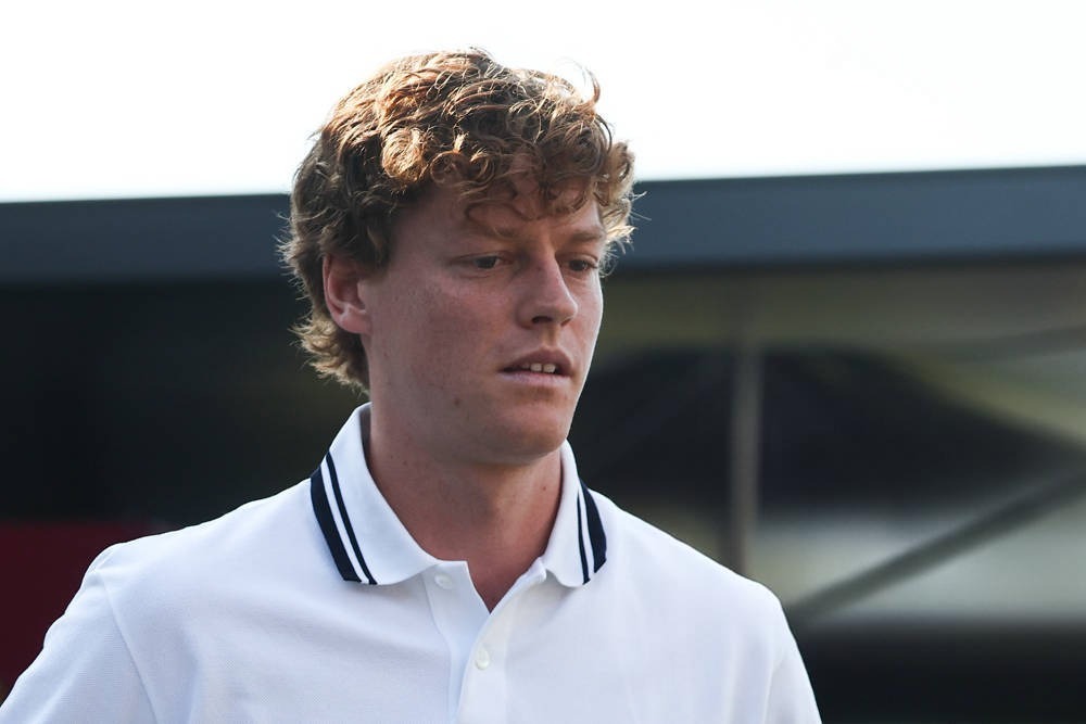 A young man with curly reddish-brown hair in a white polo with navy collar trim, looking downward in a paddock-style setting with dark structures behind him.