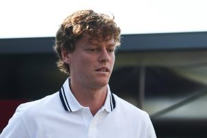 A young man with curly reddish-brown hair in a white polo with navy collar stripes, looking downward in a paddock-like setting with a dark structure behind him