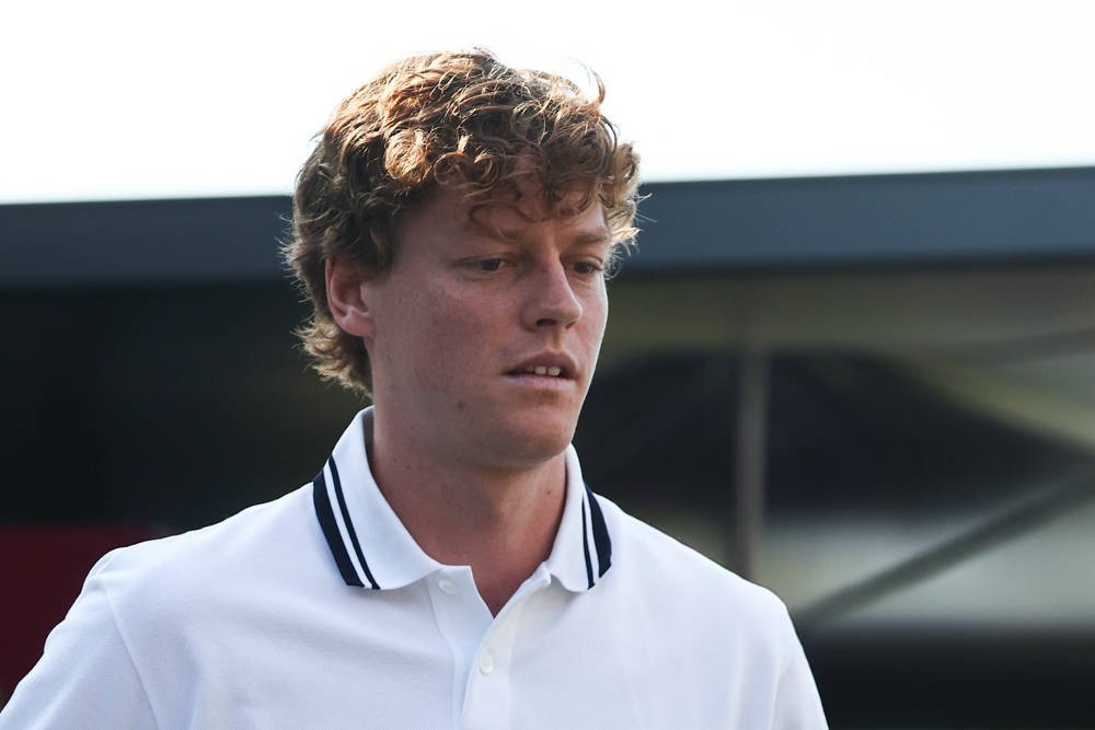 A young man with curly reddish-brown hair in a white polo with navy collar stripes, looking downward in a paddock-like setting with a dark structure behind him