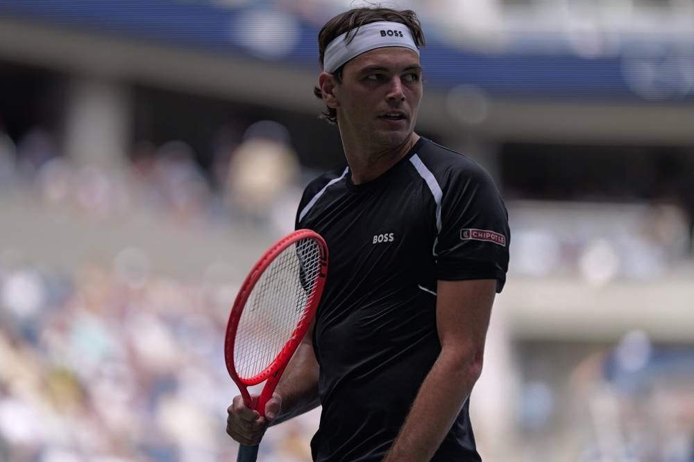 Tennis player in black BOSS shirt and white headband holding a red racket on court, as referenced in the Taylor Fritz article