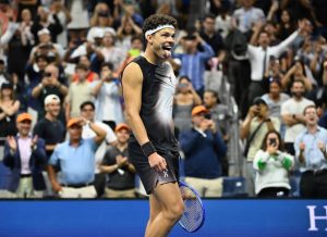 A tennis player in a black sleeveless top and white headband sticks out his tongue and celebrates on court as a crowd cheers and films at a US Open match.