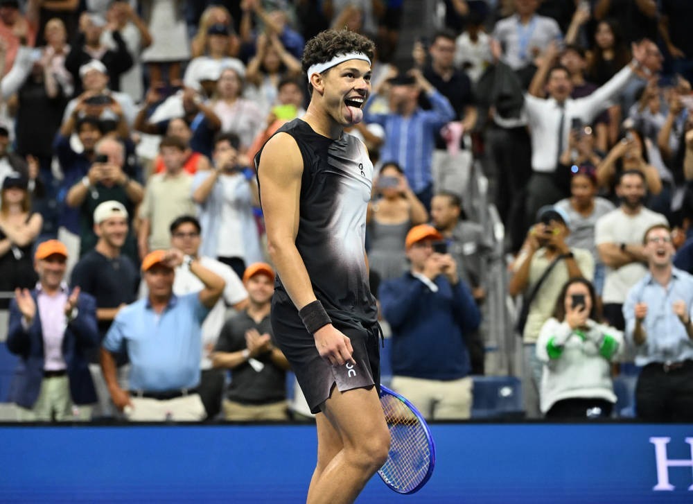 A tennis player in a black sleeveless top and white headband sticks out his tongue and celebrates on court as a crowd cheers and films at a US Open match.