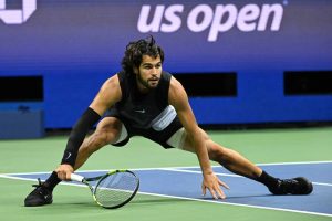 Tennis player in black Nike sleeveless top and shorts, crouched low to reach a shot on a blue-and-green court at the US Open.