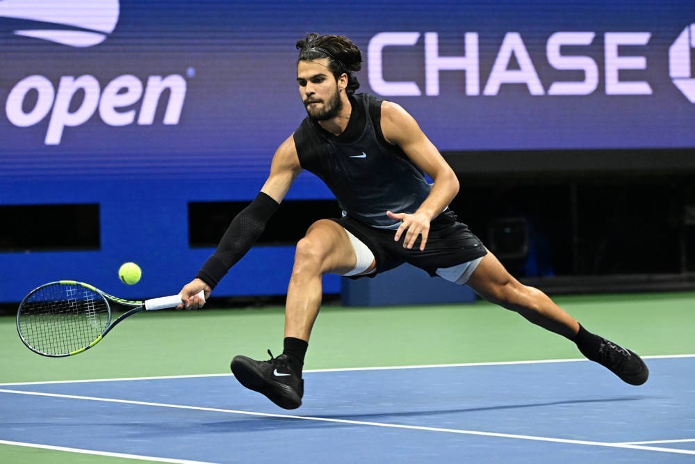 A tennis player in black sportswear lunges low to return a ball on a blue court at the US Open, with Chase signage behind him.